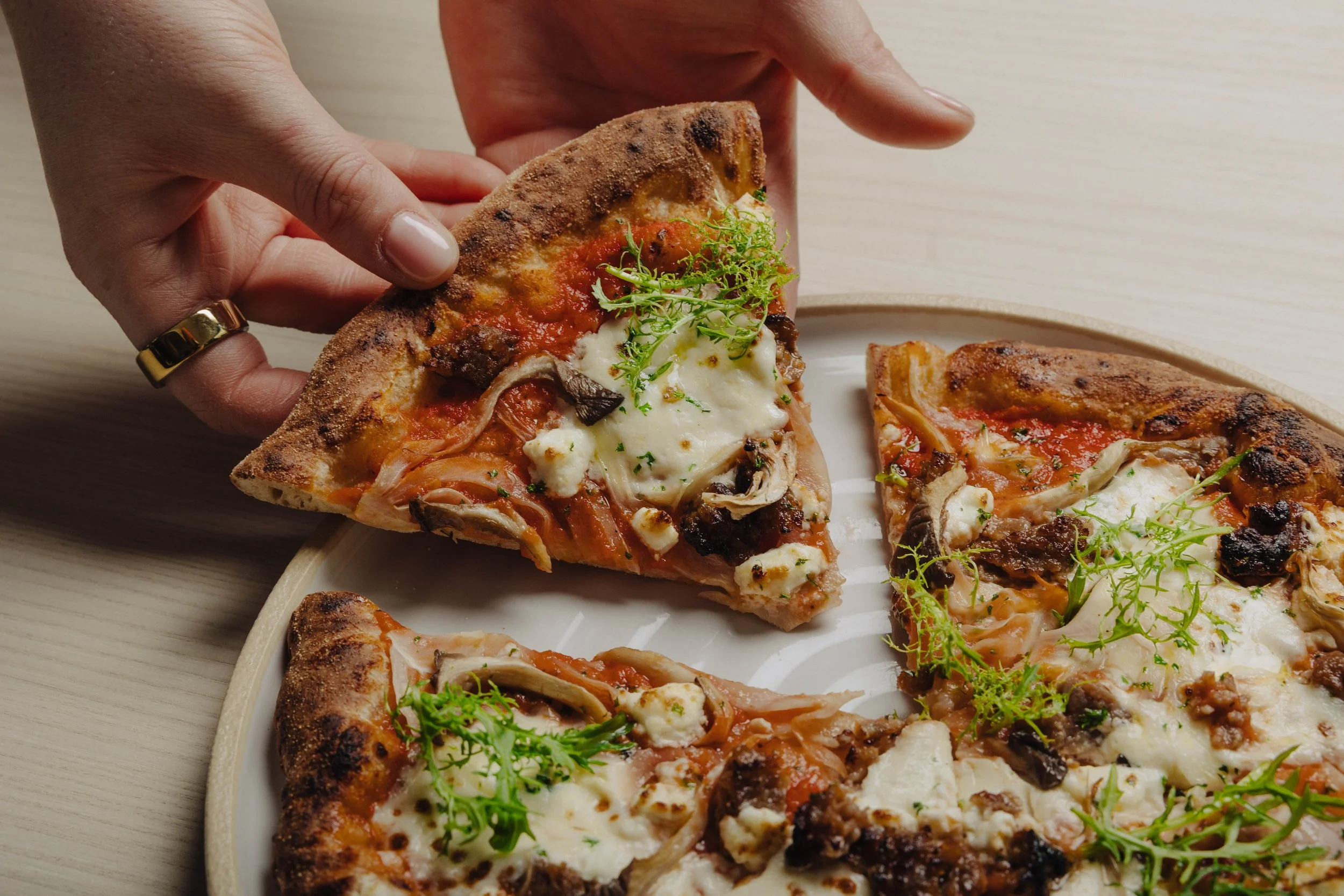 Hand holding a slice of pizza with toppings like cheese, mushrooms, and greens on a plate.