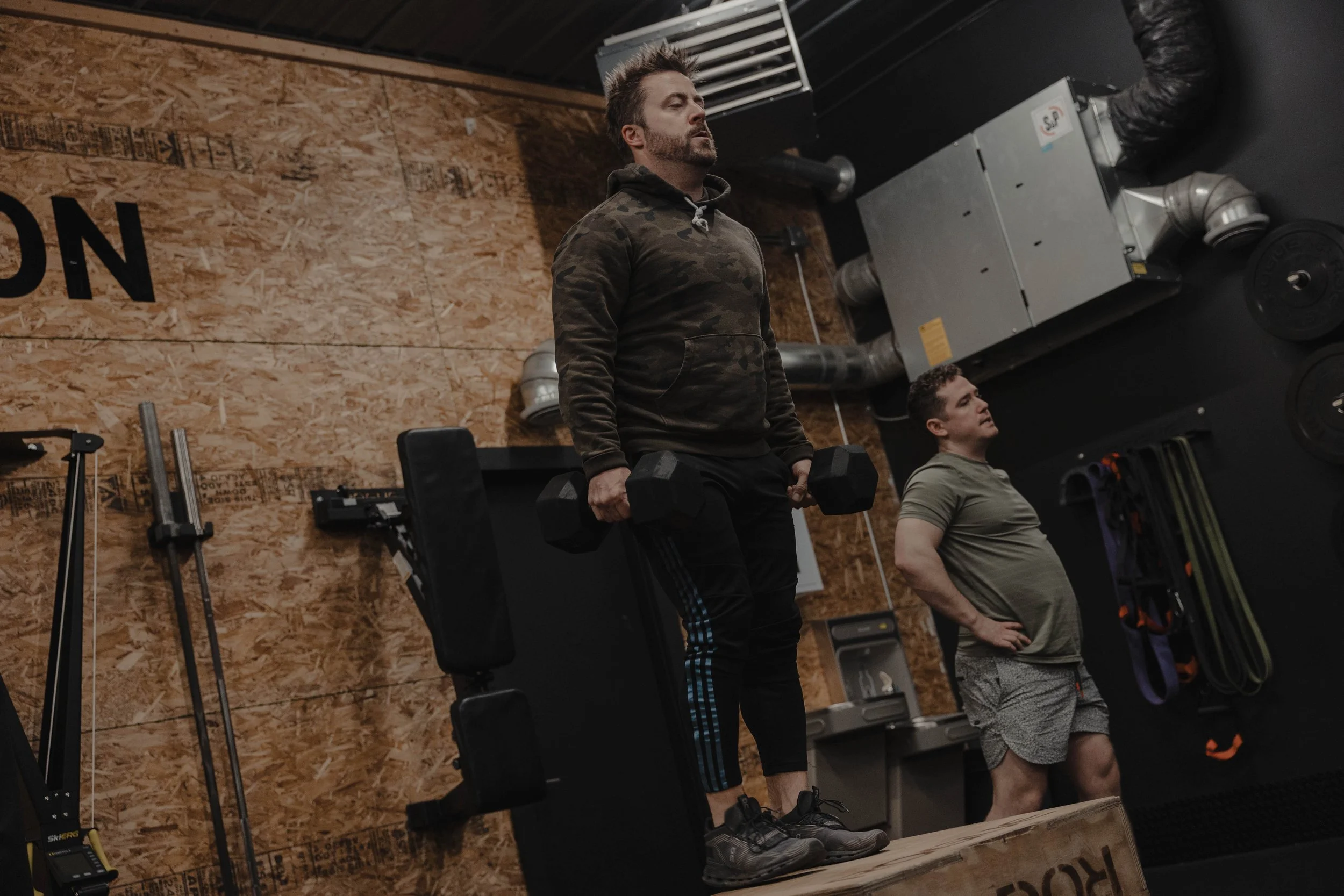 Two men in a gym, one holding dumbbells on a box and the other standing with hands on hips, against a wooden wall background.