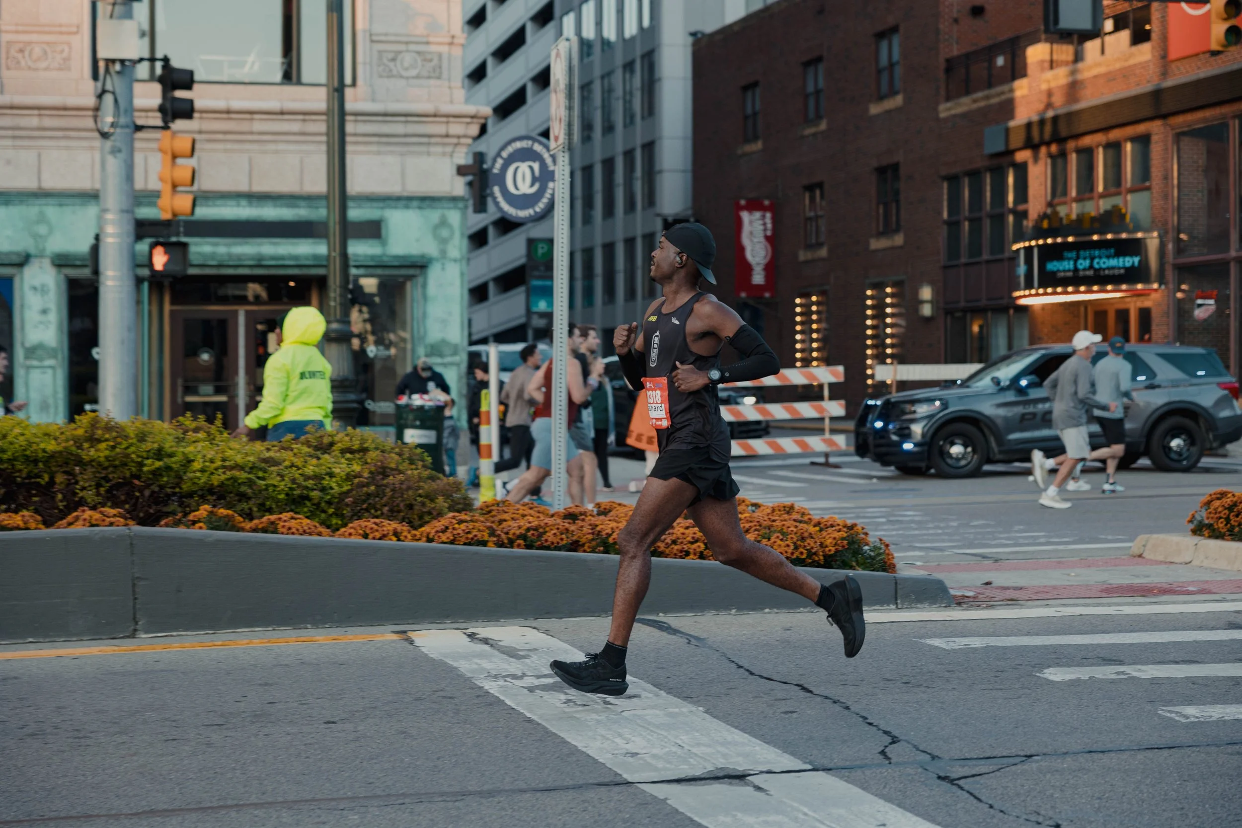 Runner in a race crossing an intersection in an urban area with a police car and comedy club in the background.