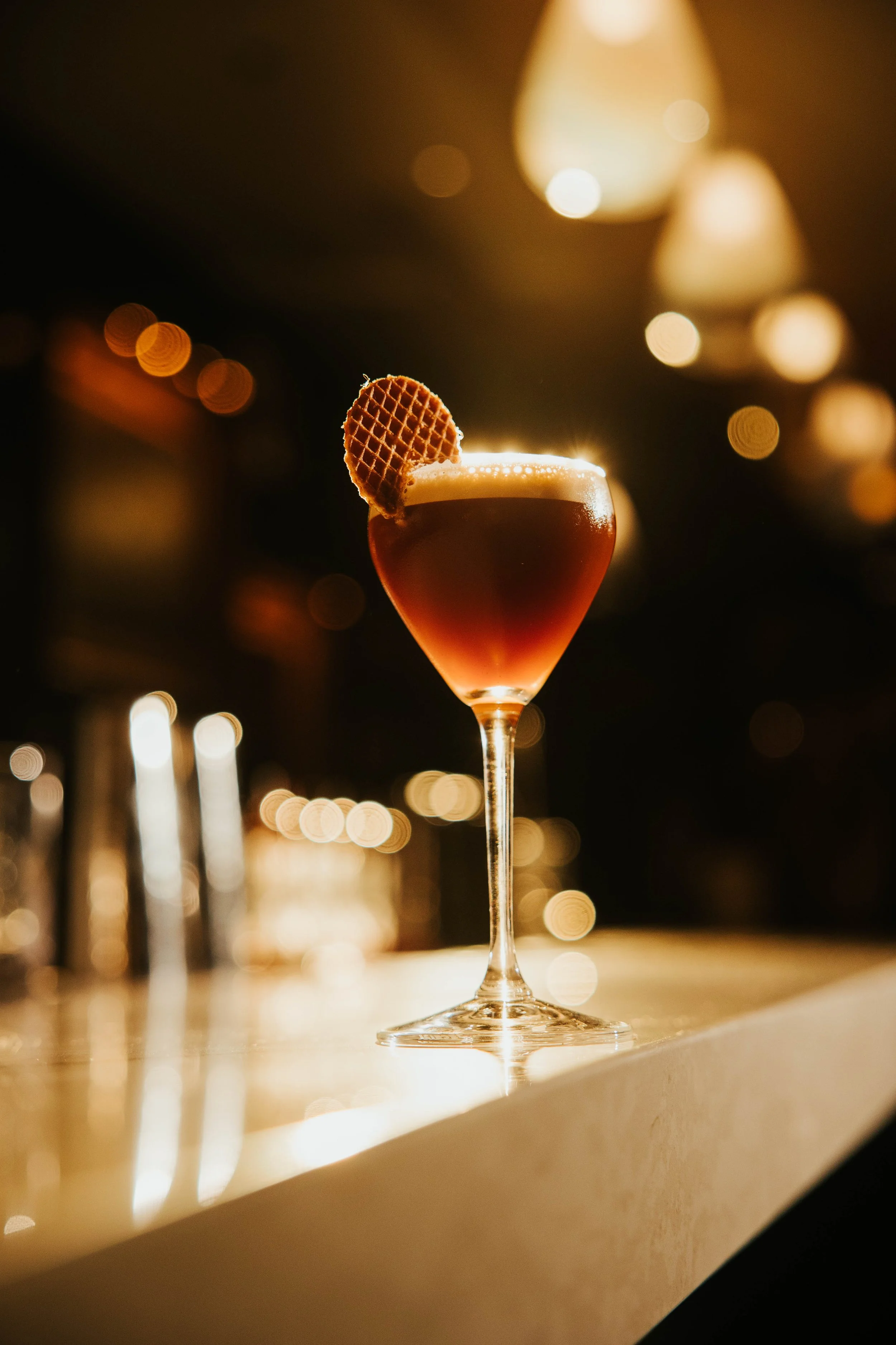 Cocktail in a glass with a waffle garnish on a bar counter with blurred lights.