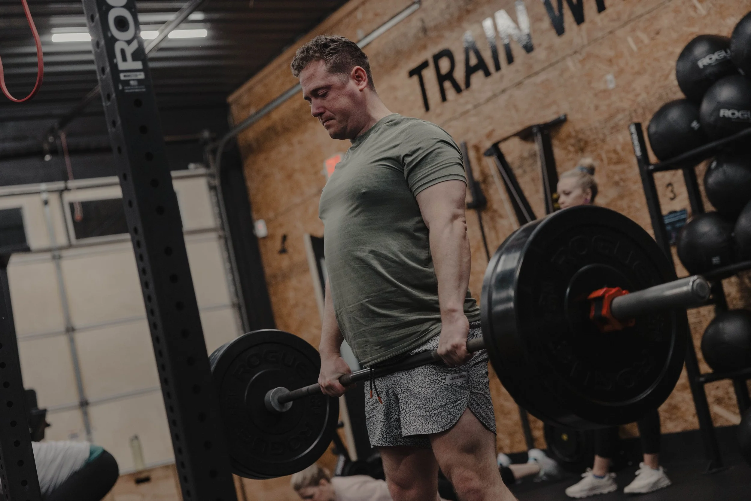 Person in a gym lifting a barbell with weight plates, wearing a green shirt and patterned shorts, with exercise equipment visible in the background.
