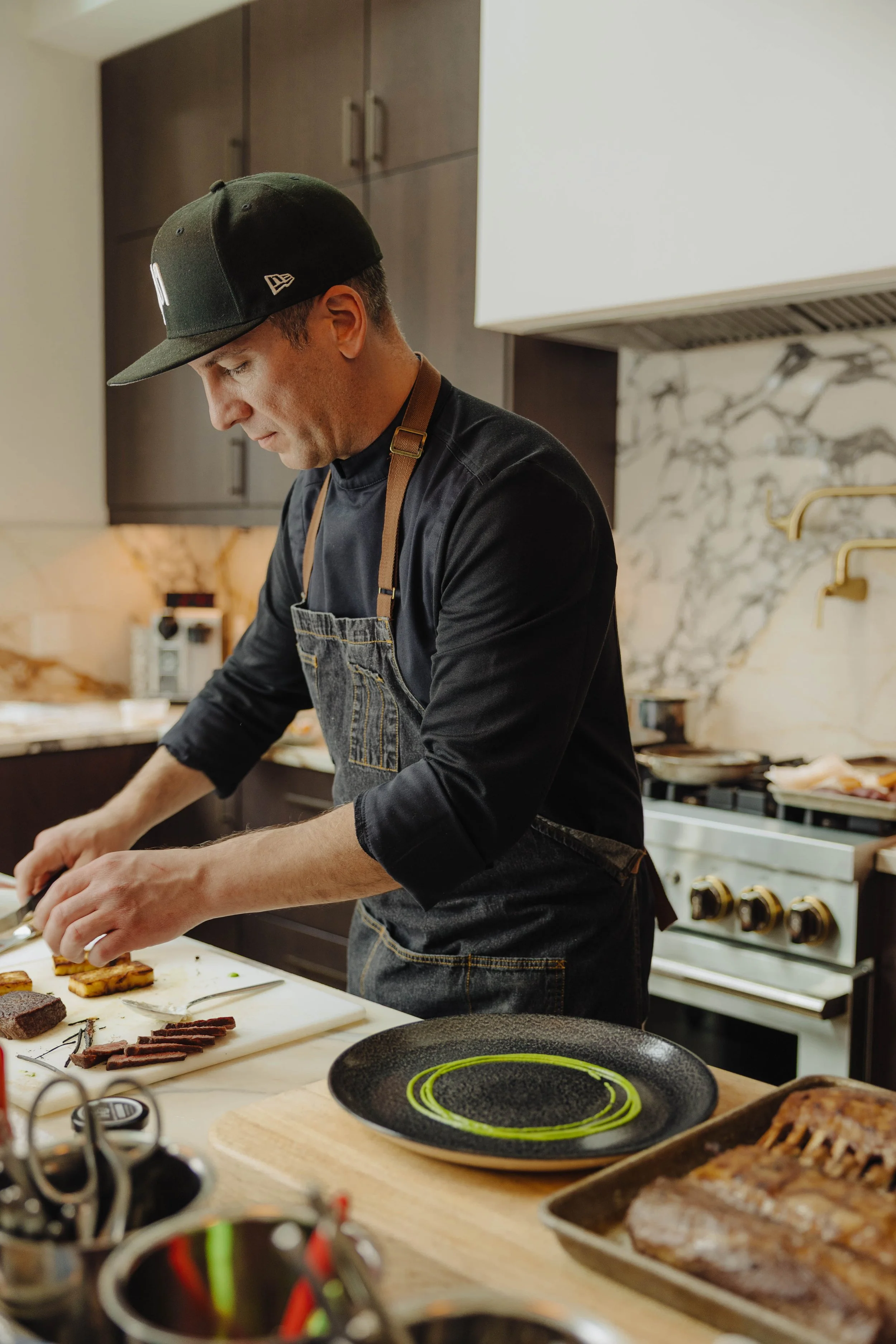 Man wearing a black cap and apron preparing food in a modern kitchen with a marble backsplash and professional-grade stove.