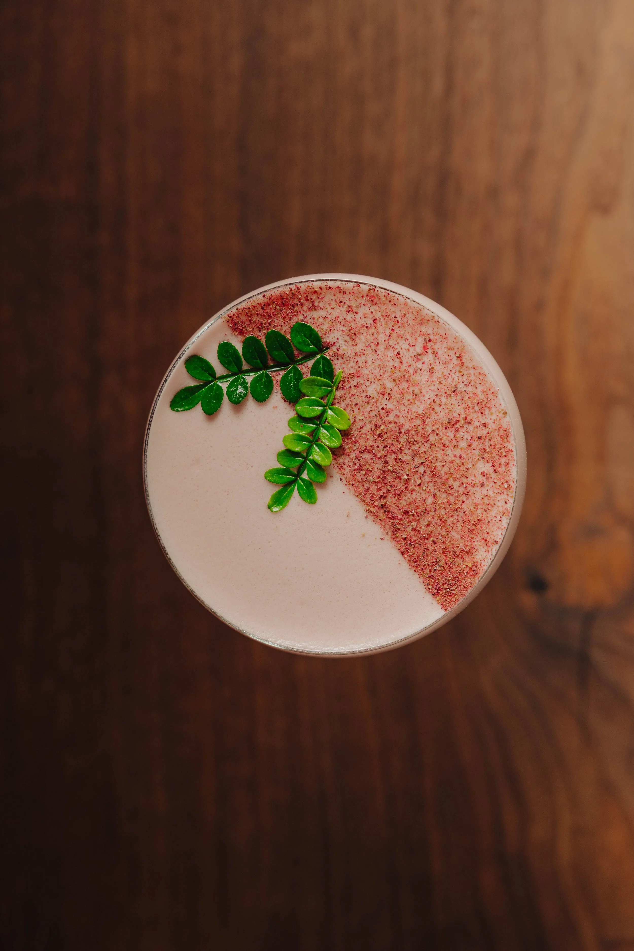 A creamy cocktail with a frothy top, garnished with green leaves and red powder, served on a wooden table.
