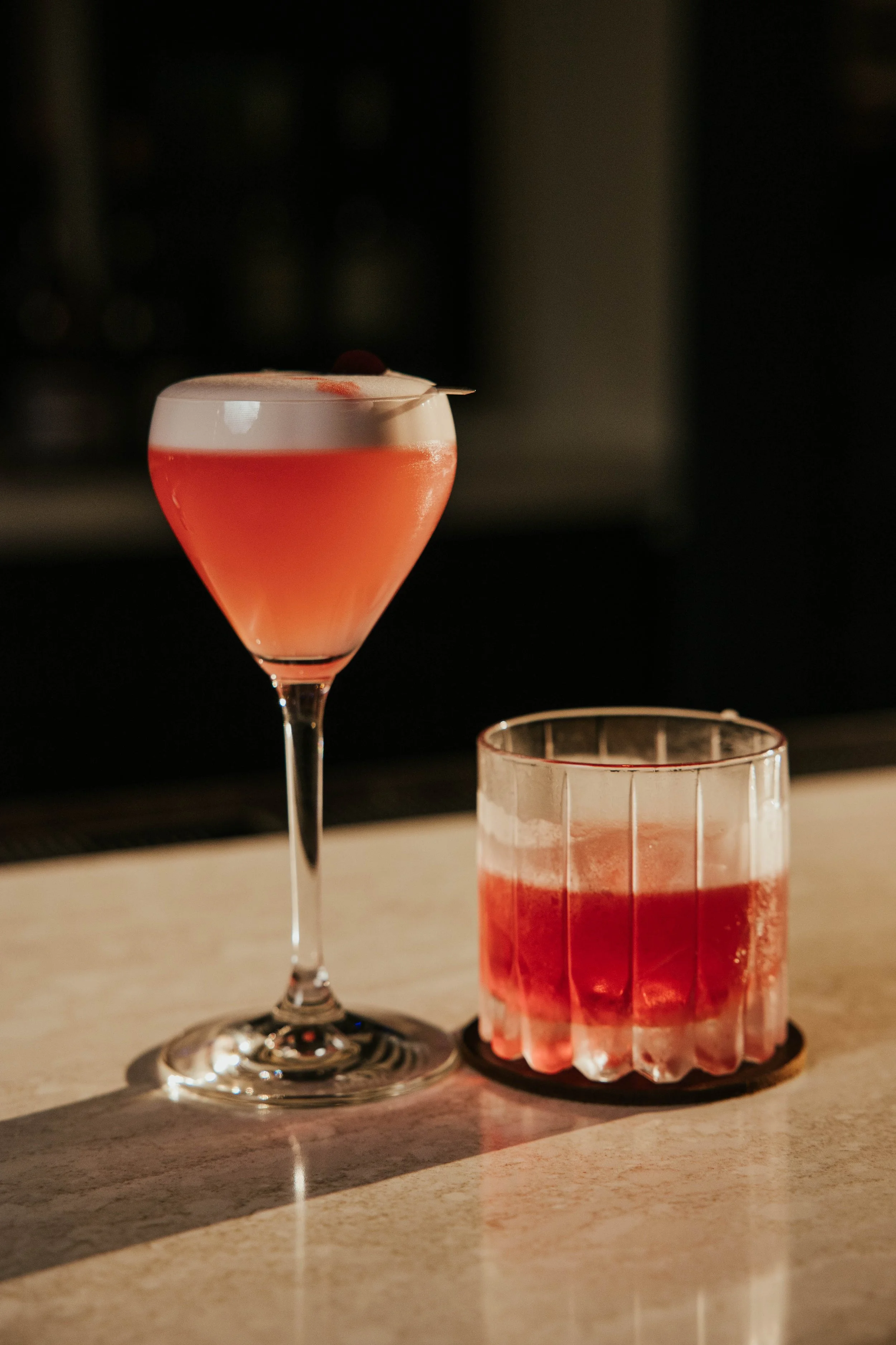 Two red mixed drinks; one in a tall stemmed glass garnished with a cherry, and the other in a short tumbler with ice on a marble surface.