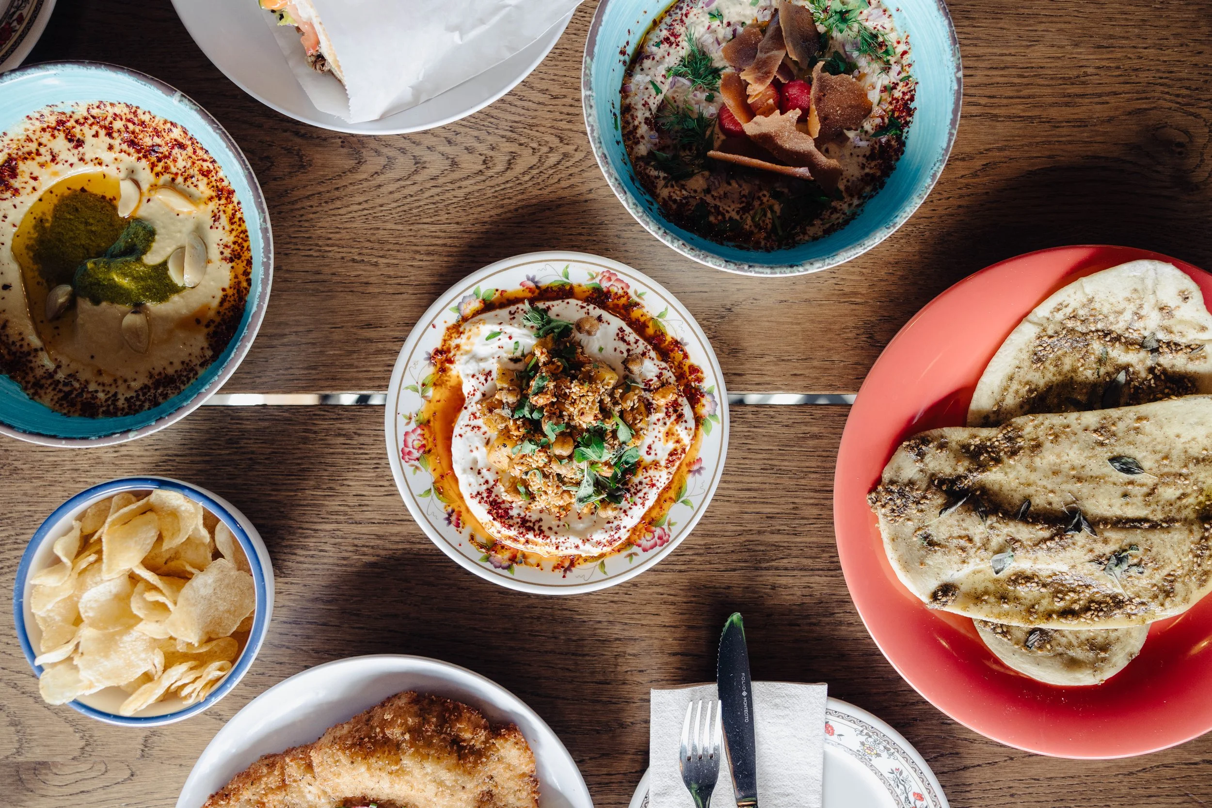 Assorted Middle Eastern dishes including hummus, flatbread, chips, and a yogurt-based dish on a wooden table.