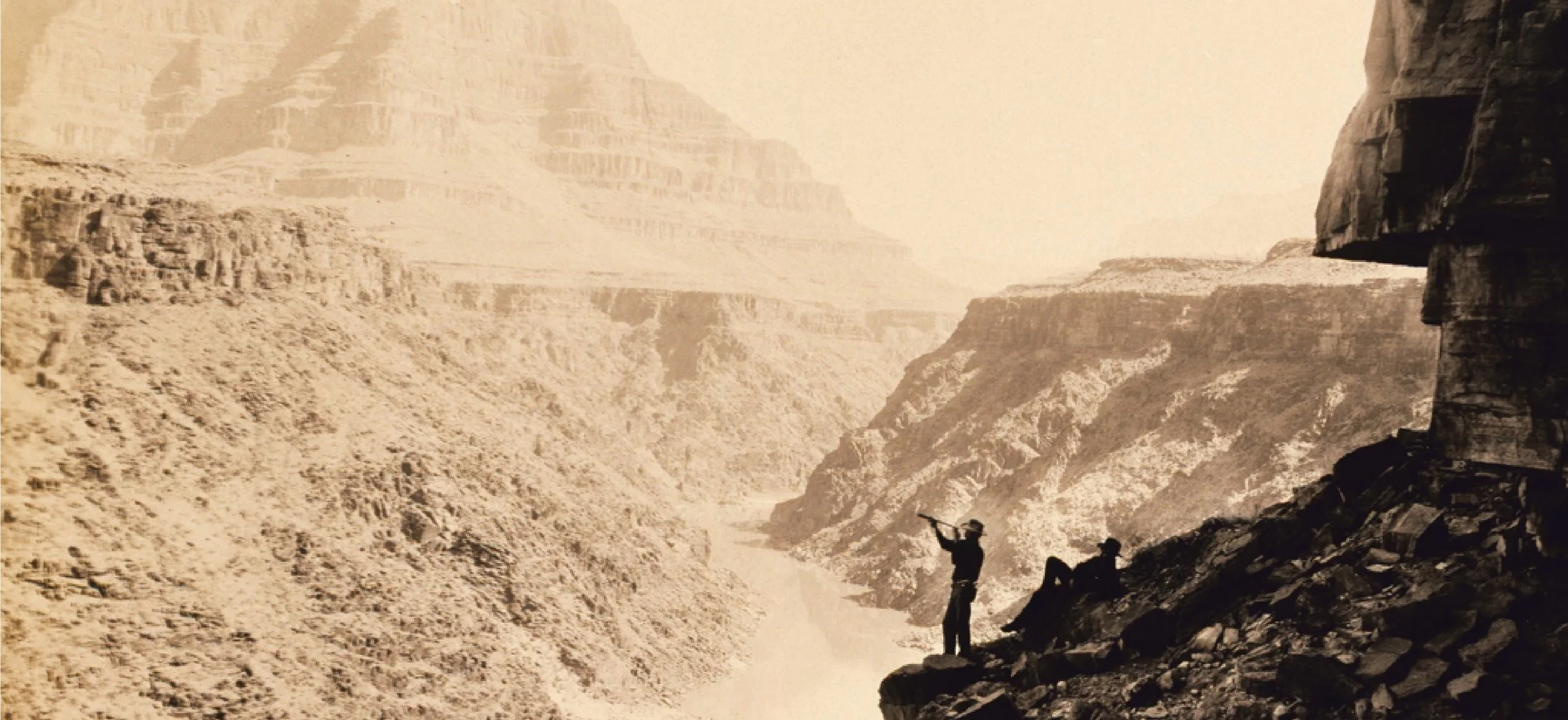 Silhouette of two people on rocky ledge near the Grand Canyon with a river below, sepia tone image.