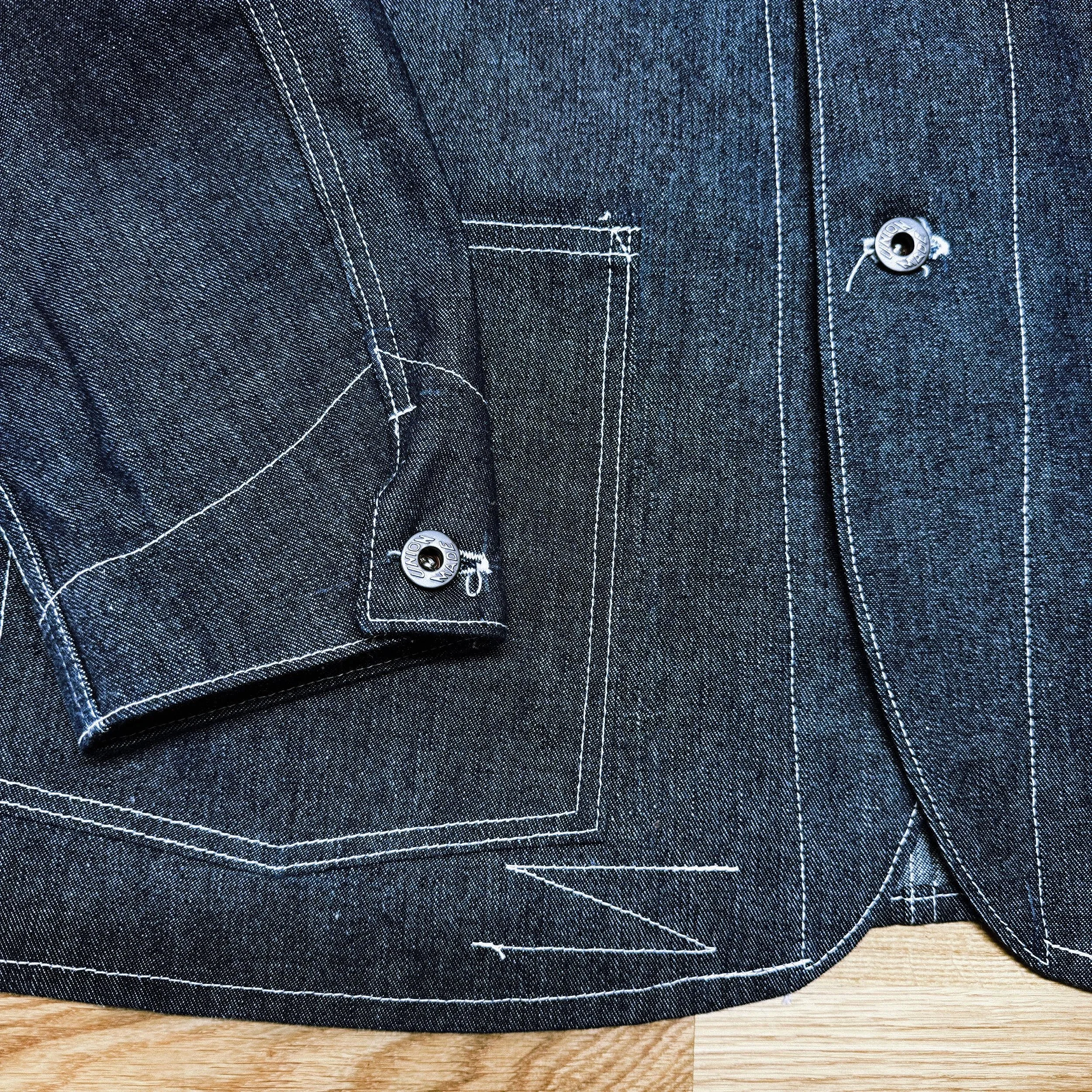 Close-up image the bottom of an early 1900’s inspired denim chore coat with white contrast stitching, featuring part of a pocket, a cuff with a metal button, and hem.