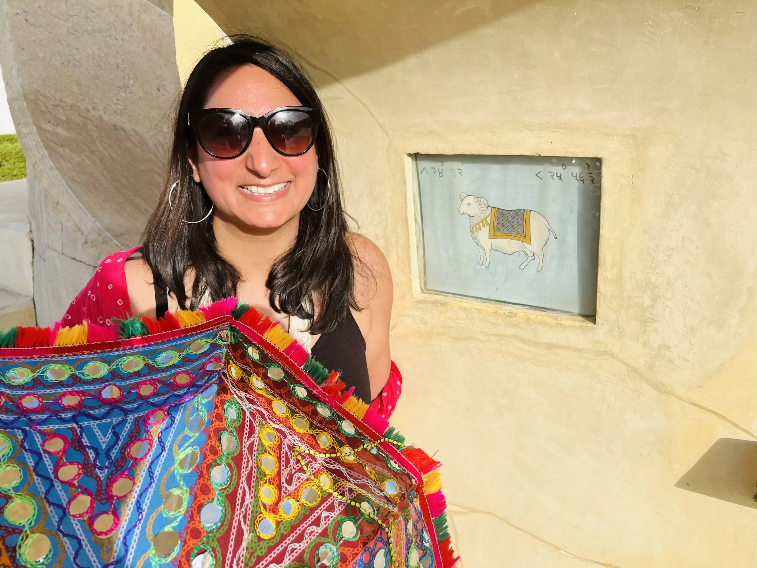 A woman with long dark hair wearing sunglasses and a red dress with white polka dots, holding a colorful woven textile in front of a yellow wall with a small window depicting a goat.