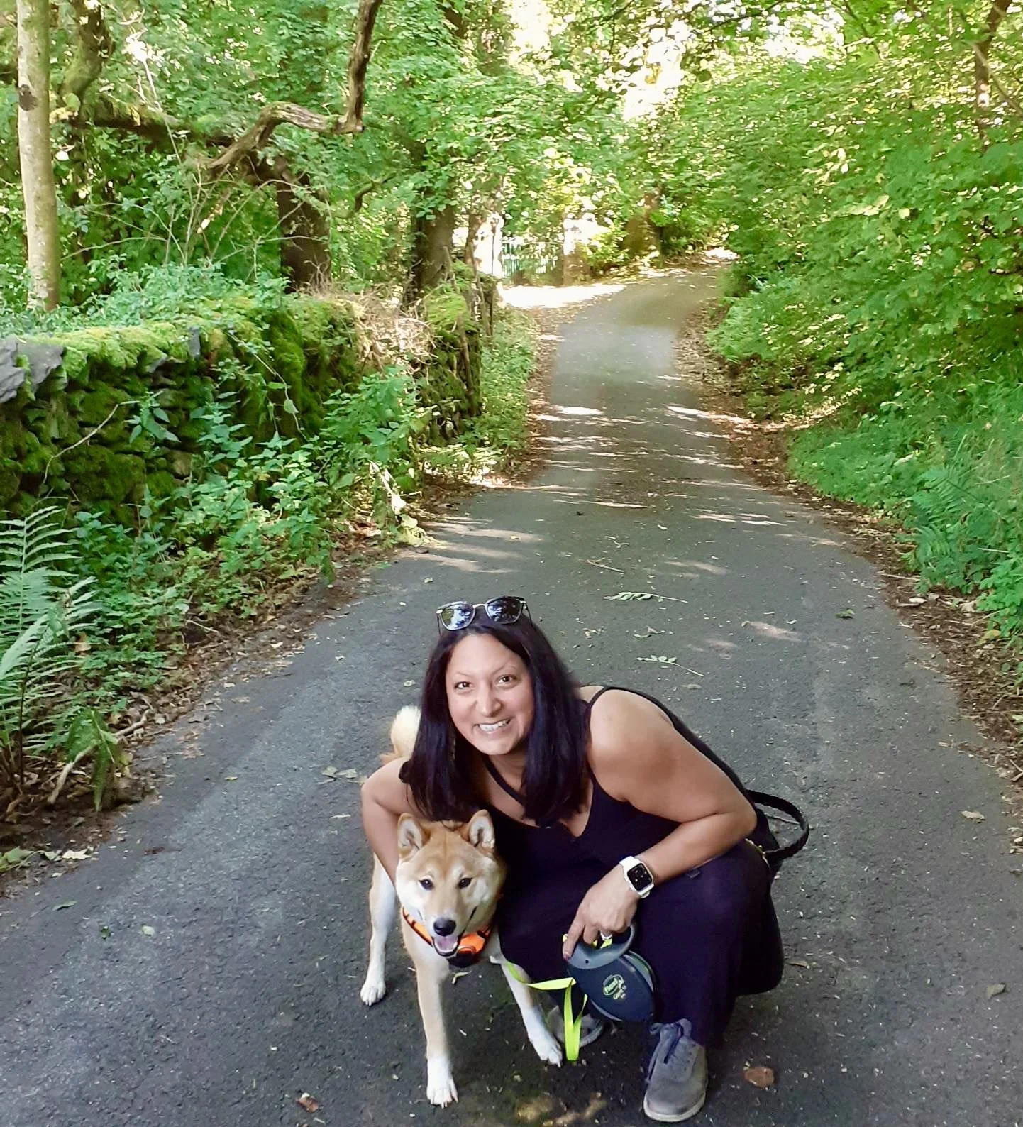 A woman with long dark hair, wearing a black tank top and black pants, is kneeling on a paved trail in a green, wooded area, smiling and holding a Shiba Inu dog with a harness.