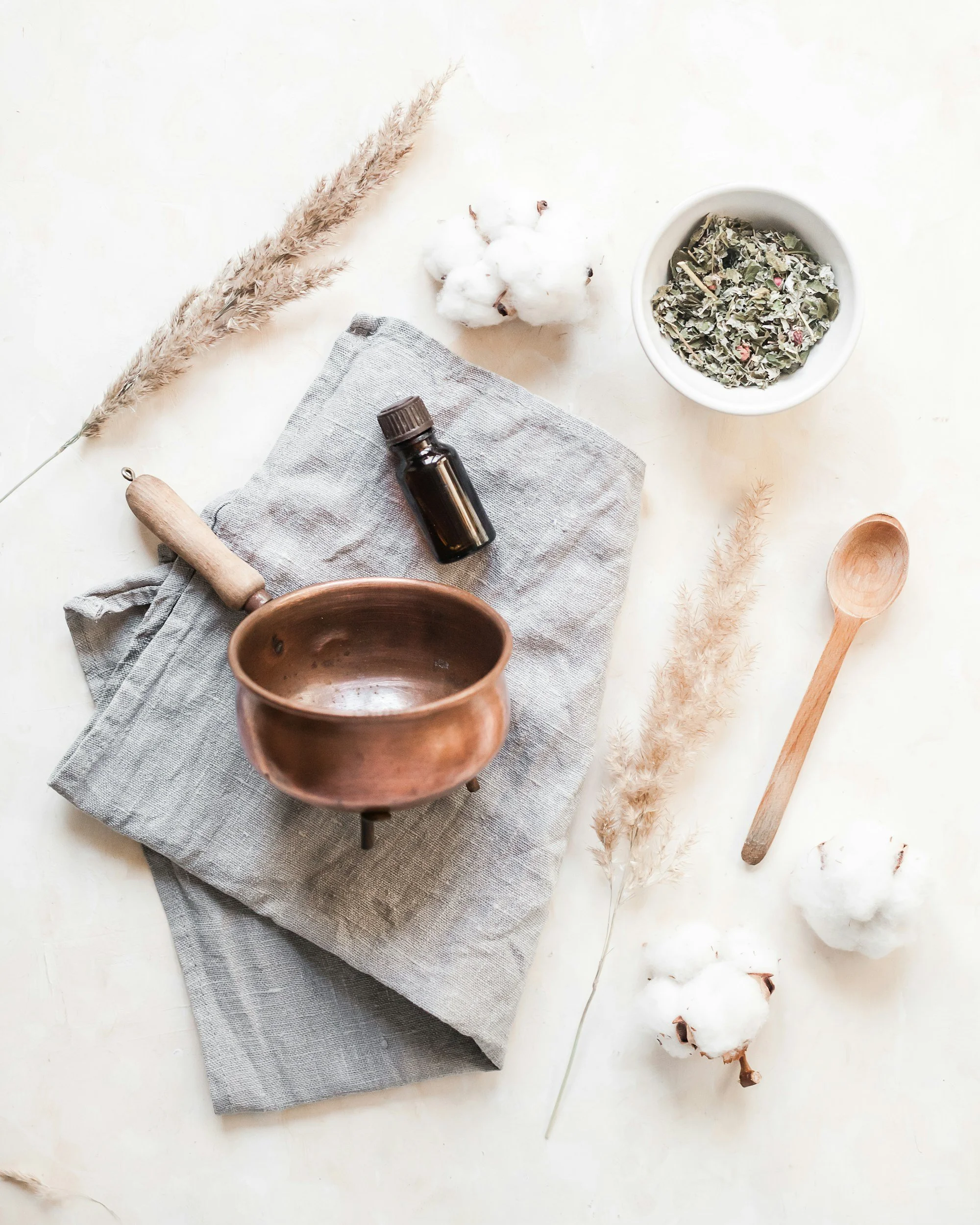 Flat lay of natural and wellness items including a cotton branch, dried herbs in a white bowl, an amber essential oil bottle, a gray linen cloth, a wooden spoon, and a wooden pestle on a white surface.