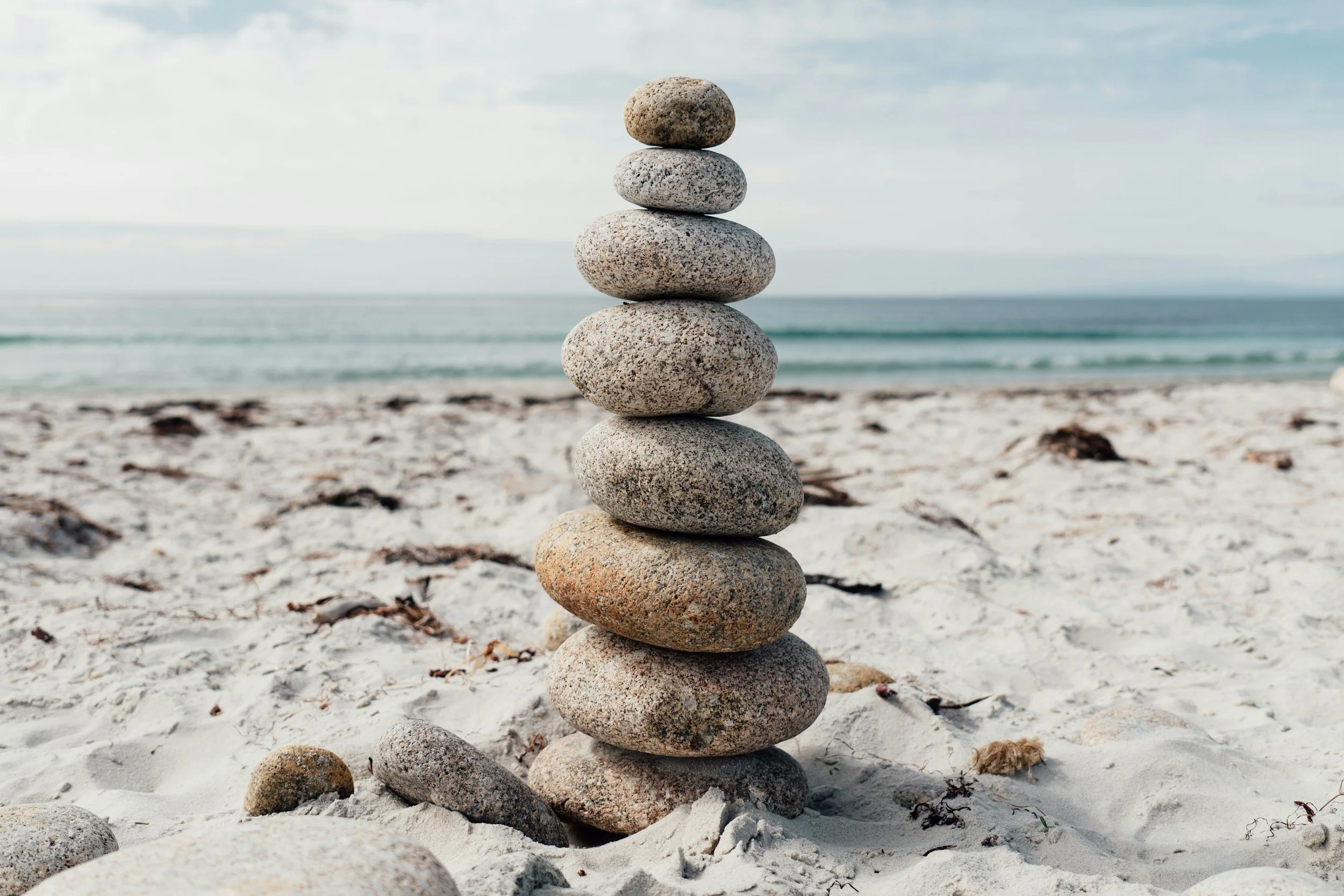 Stack of seven gray and beige smooth stones balanced vertically on a sandy beach with the ocean and cloudy sky in the background.