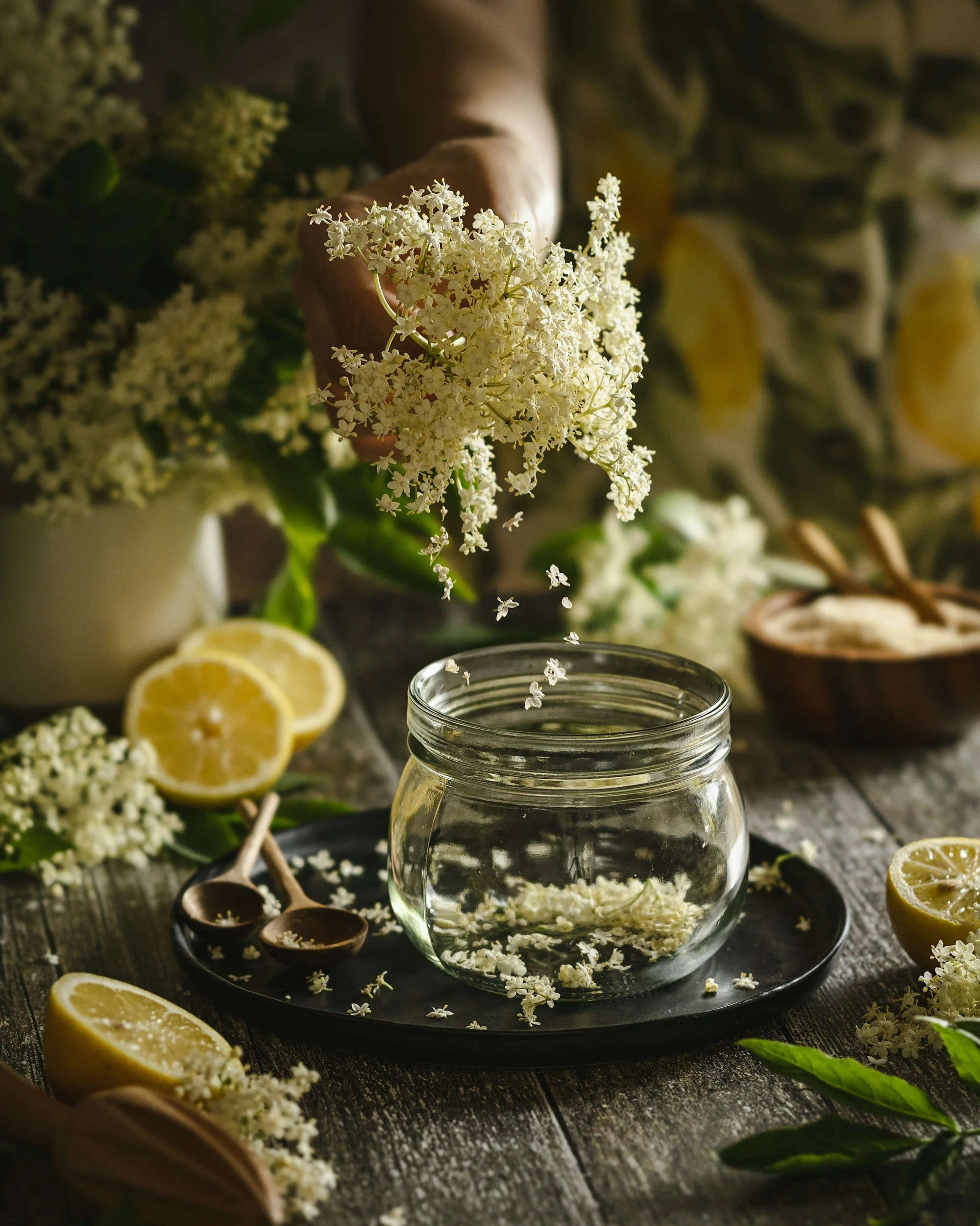 A jar of water with white elderflower blossoms inside, surrounded by lemon slices, elderflower sprigs, and small wooden spoons on a dark rustic tray. A person is dropping elderflower blossoms into the jar, with additional elderflower and lemon slices in the background.