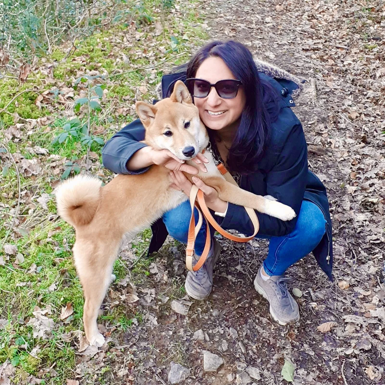 A woman wearing sunglasses, a dark jacket, and blue jeans crouches on a leaf-covered trail in a forest, holding a tan and white Shiba Inu dog with a curled tail and a collar with a leash.
