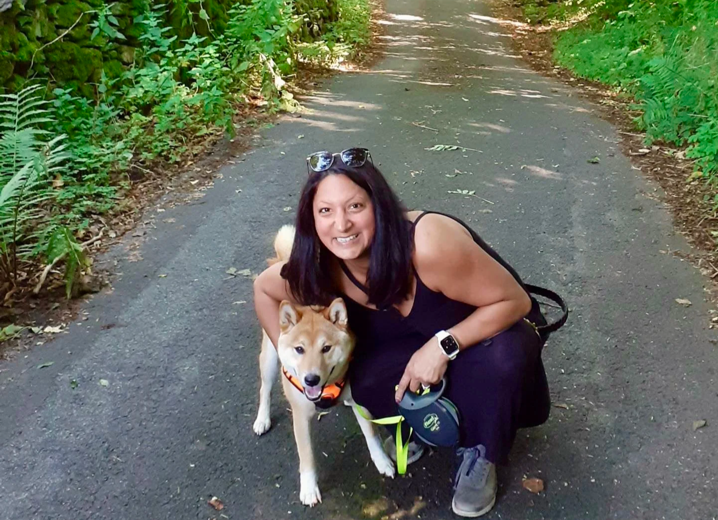 A woman kneeling on an outdoor trail with a friendly dog in a forested area.