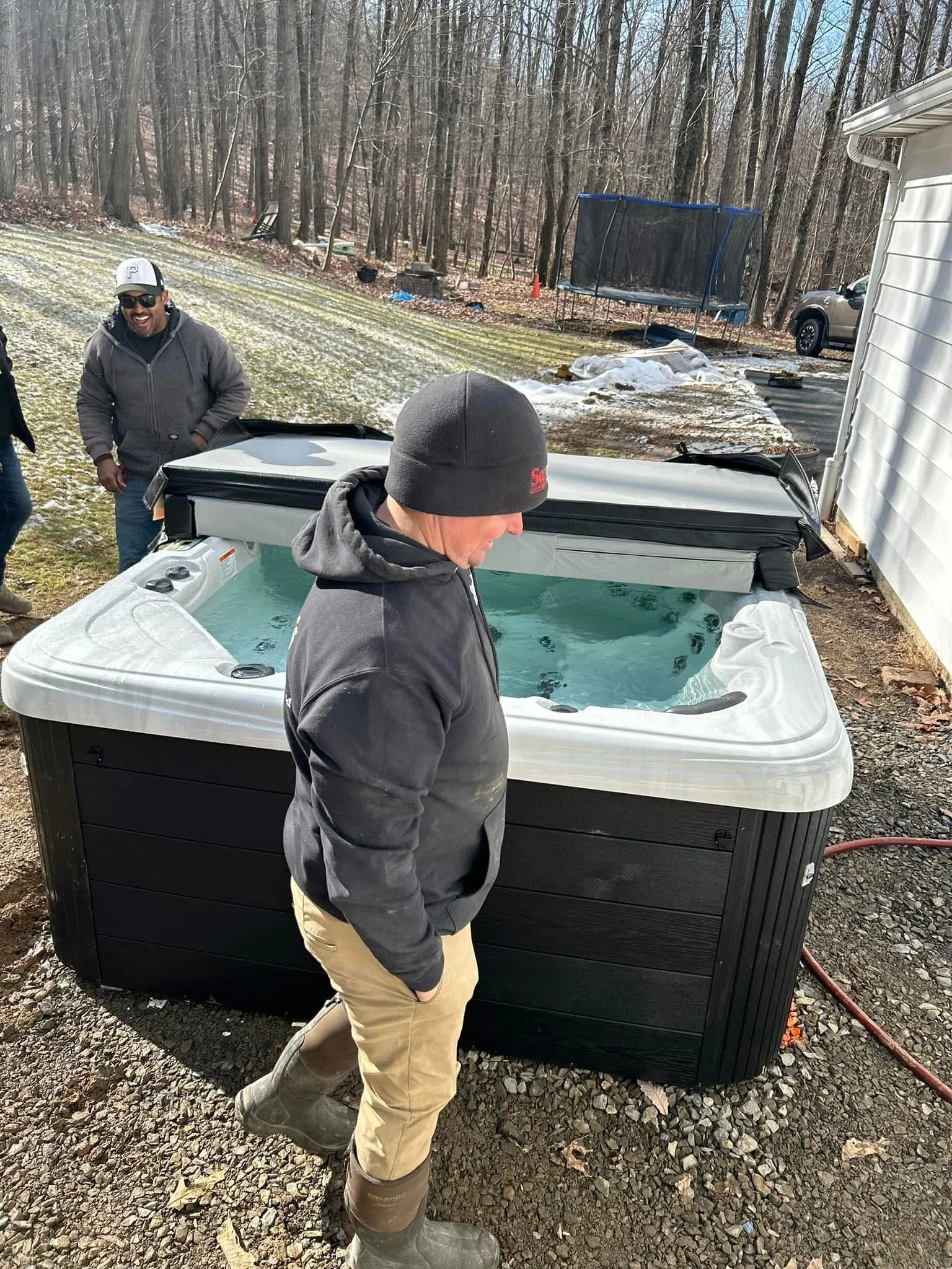 Two people near a hot tub outdoors with trees and a trampoline in the background.
