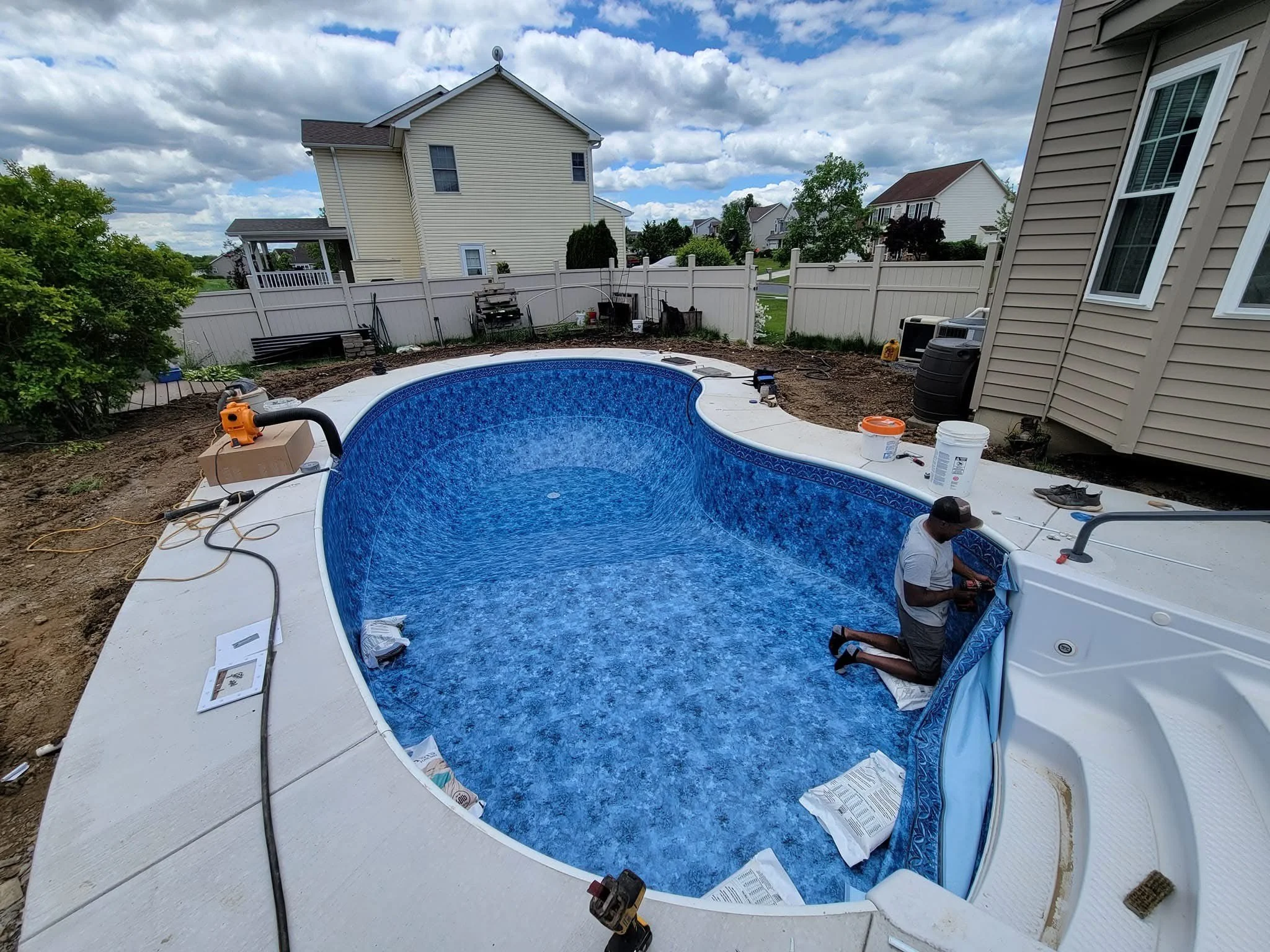 A worker installing a blue liner in an empty swimming pool, with surrounding residential homes in the background. Construction materials and tools are visible around the pool.
