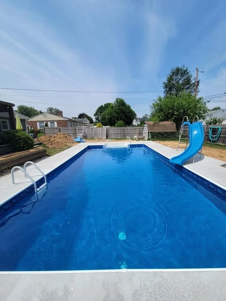 Backyard swimming pool with a slide and ladder, surrounded by a fence and trees, under a clear blue sky.