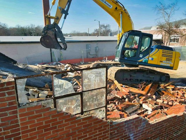 Excavator demolishing a brick building with broken windows and debris.