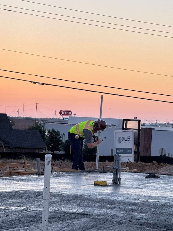 A construction worker in a yellow safety vest and helmet is working on a construction site during sunset. He is bent over, measuring or marking the freshly laid concrete slab, with construction signage and buildings visible in the background.