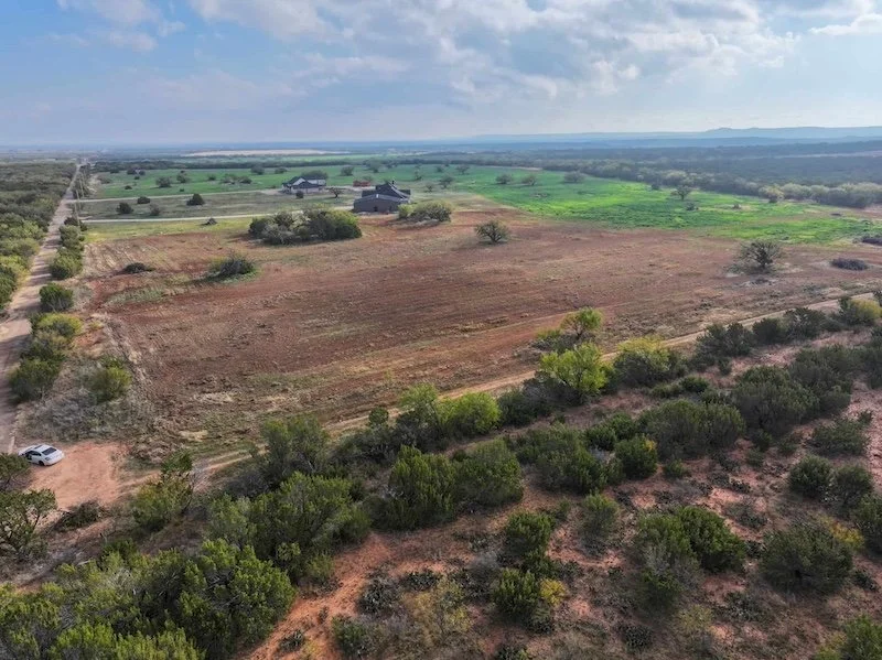Aerial view of rural landscape with open fields, scattered trees, and distant buildings under a cloudy sky.