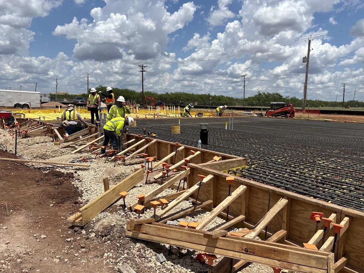 Construction workers in safety vests and helmets working on a concrete foundation with rebar reinforcement, with a bright sky and scattered clouds overhead.