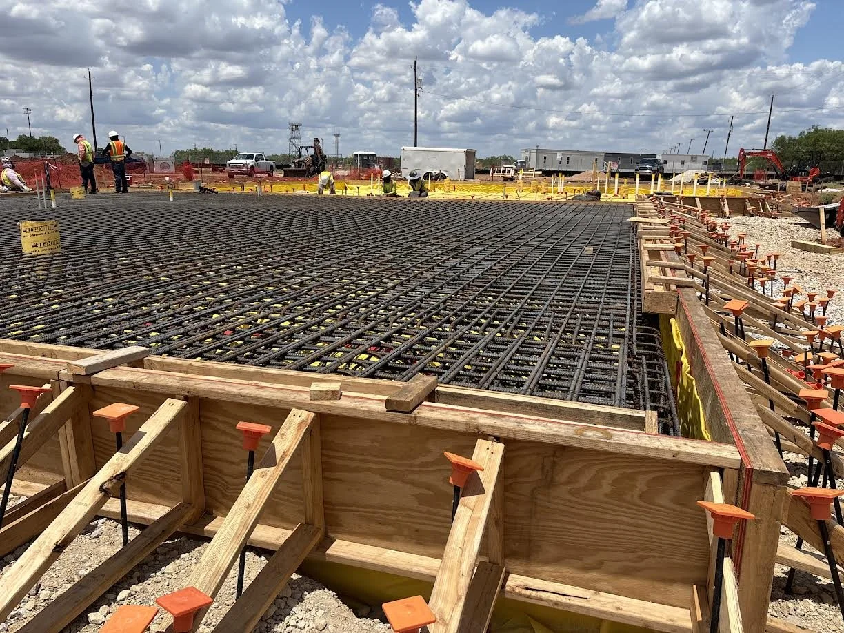 Construction site with rebar reinforcement for concrete slab, workers in safety vests and helmets, and construction equipment under a partly cloudy sky.