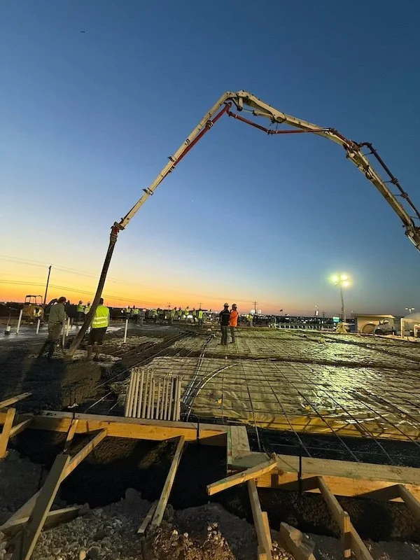Construction workers pouring concrete on a building site at sunset, with construction equipment and a clear evening sky.