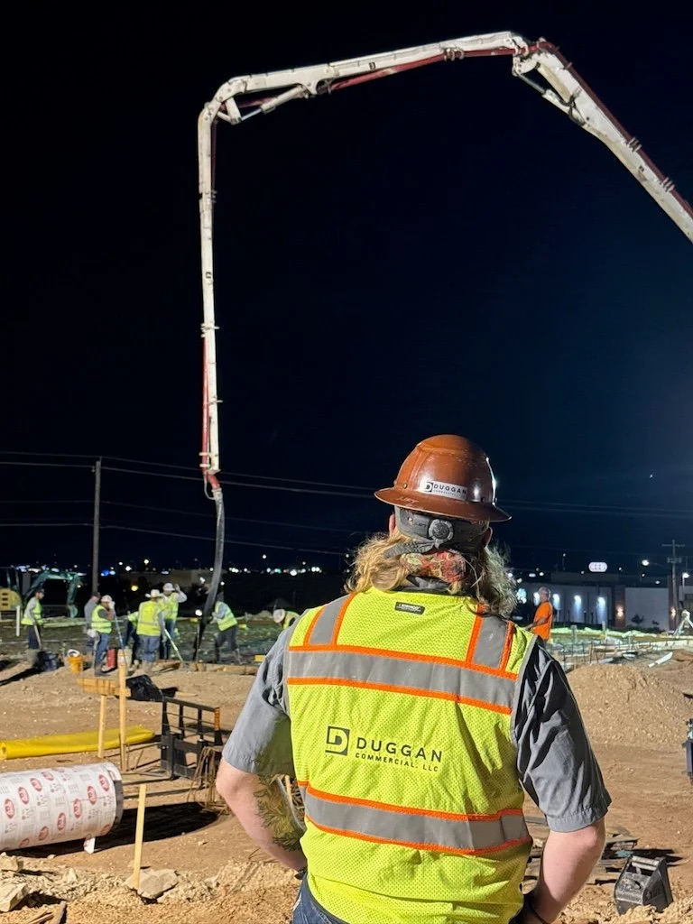 A construction worker with long hair, wearing a brown hard hat and a yellow safety vest labeled 'Duggan Commercial LLC,' observes a nighttime construction site where multiple workers are operating heavy machinery and equipment.