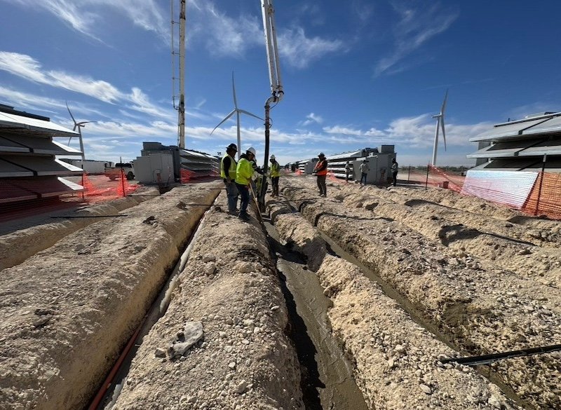 Construction site with workers pouring concrete into trenches, wind turbines in the background, and construction materials on site.
