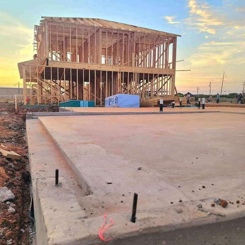 House under construction with wooden framing on a concrete foundation, against a clear sky at sunset.