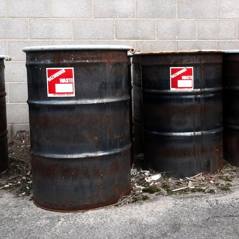 Rusty metal barrels marked with hazardous waste labels against a concrete wall.