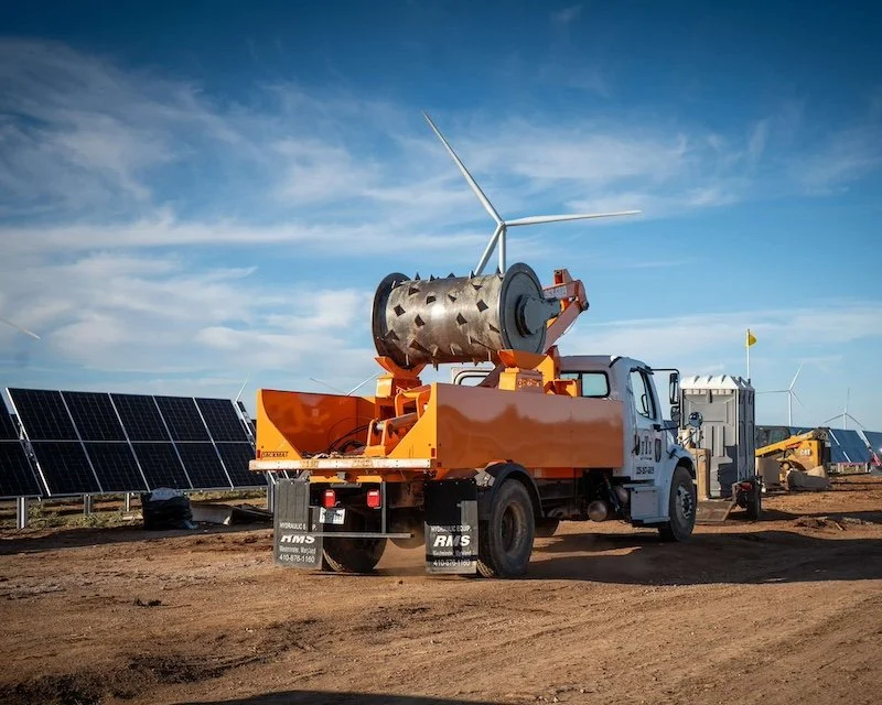 Truck with construction equipment at a renewable energy site with solar panels and wind turbines.
