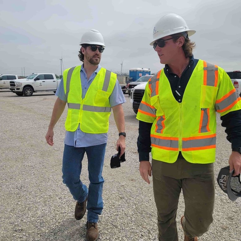 Two construction workers wearing reflective safety vests and hard hats talking at a work site with trucks and equipment in the background.