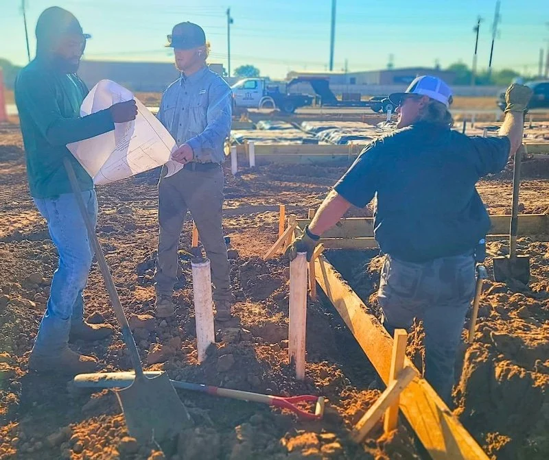 Three construction workers examining blueprints at a construction site with shovels and wooden stakes in the foreground.