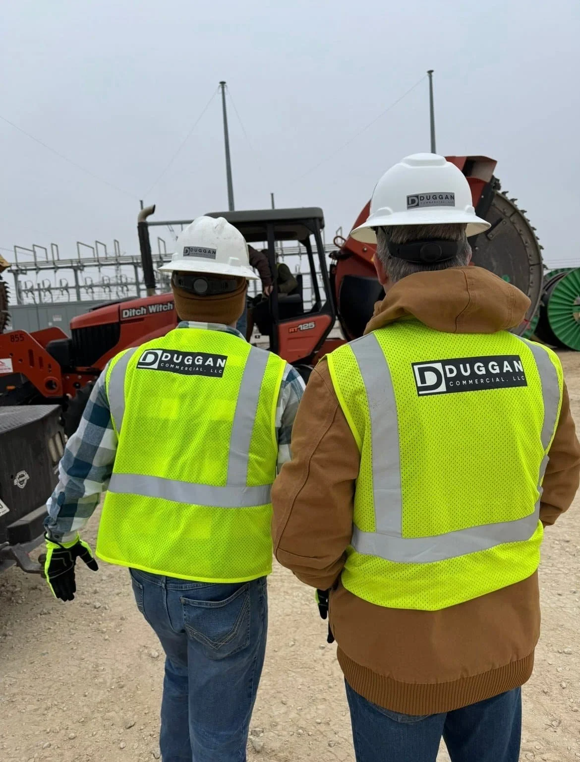 Two construction workers wearing safety vests and helmets, standing in front of a large orange construction vehicle at a work site.