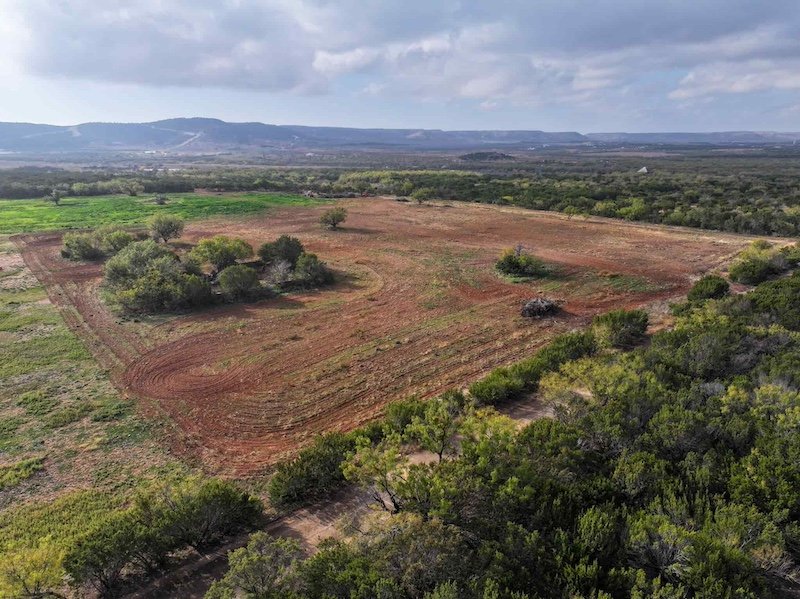 Aerial view of rural landscape with fields, scattered trees, and distant hills under a cloudy sky.