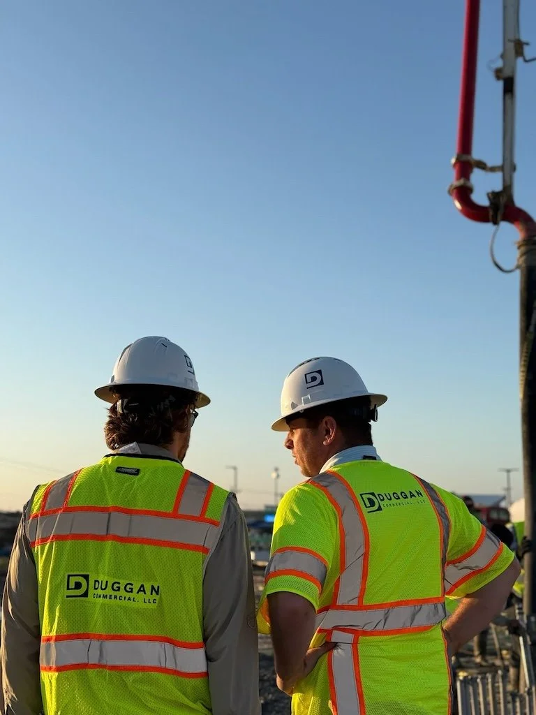 Two construction workers in yellow safety vests and white helmets standing outdoors, talking, with a clear blue sky in the background.