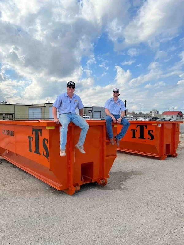 Two men sitting on orange dumpsters with blue sky and clouds in the background.
