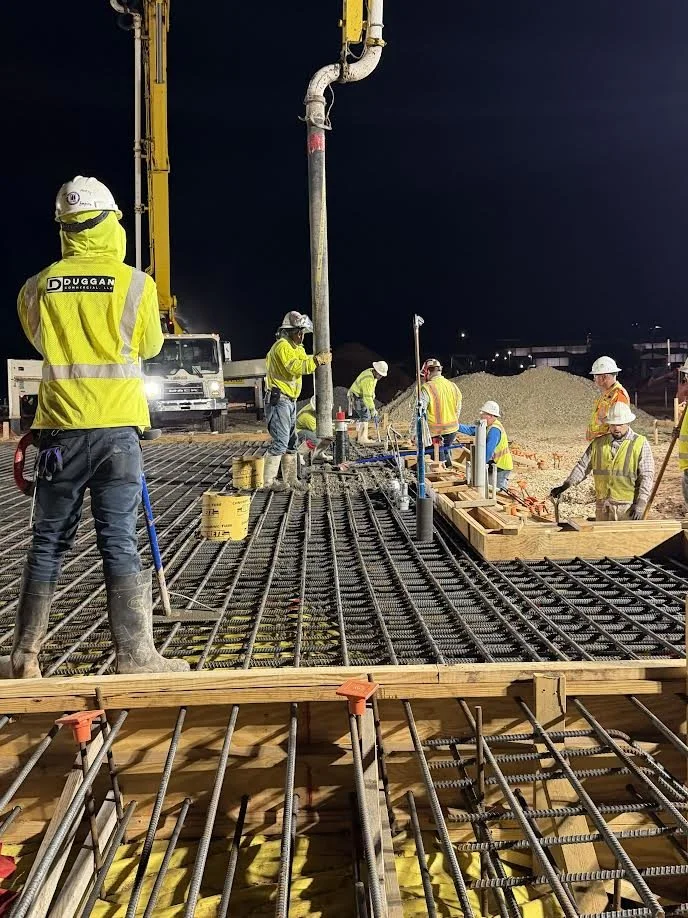 Construction workers wearing safety vests and helmets work on a bridge or elevated structure at night, with rebar and wooden formwork in place.