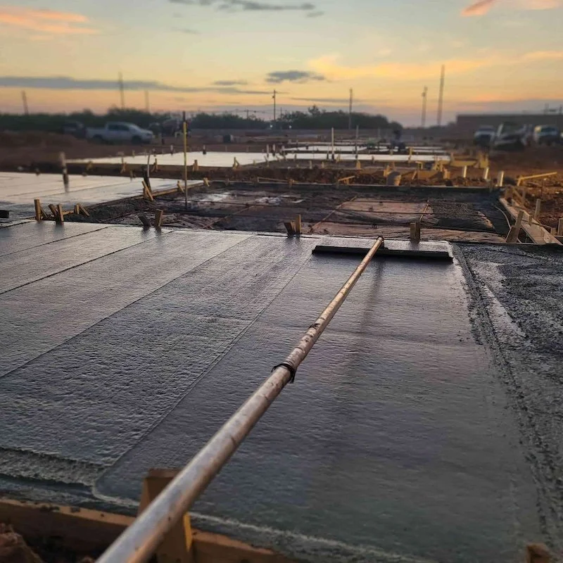 Freshly poured concrete slabs being smoothed at a construction site during sunset, with tools and wooden frames visible.