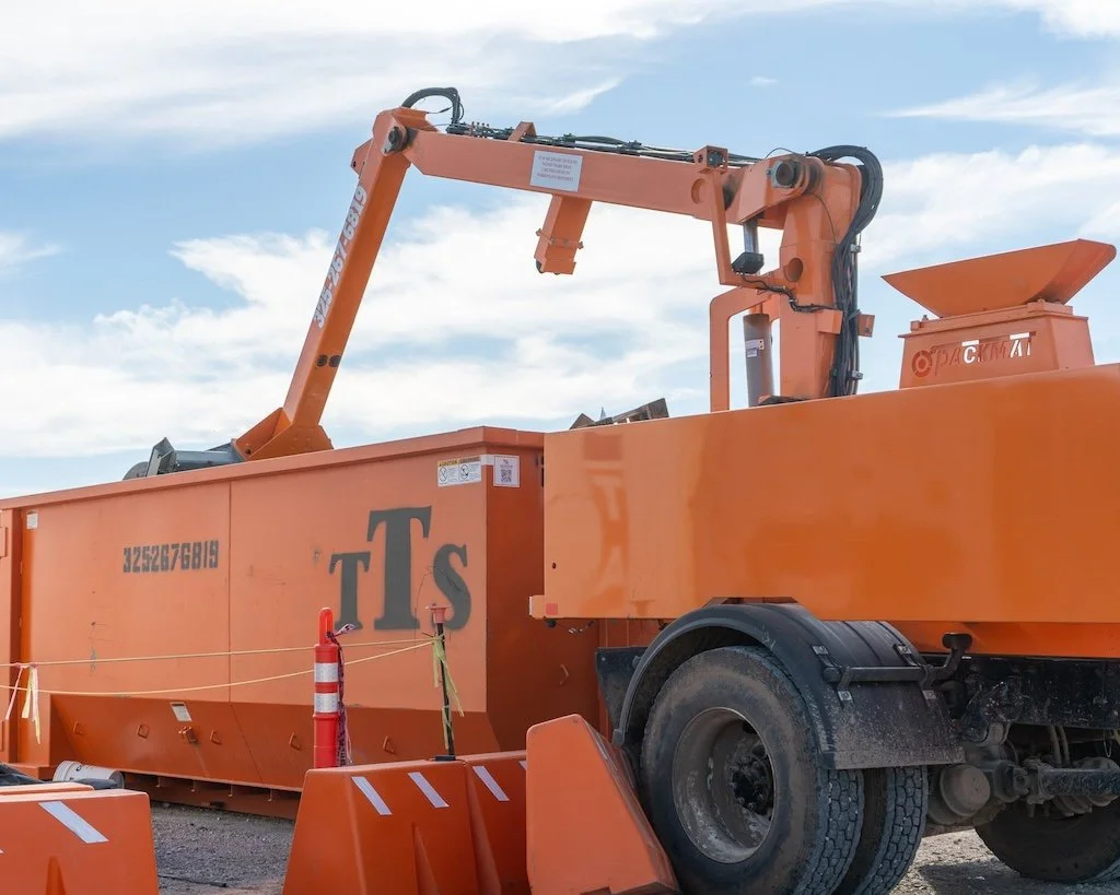 An orange industrial compactor with a crane arm, labeled "PACKMAT," on a construction site against a blue sky.