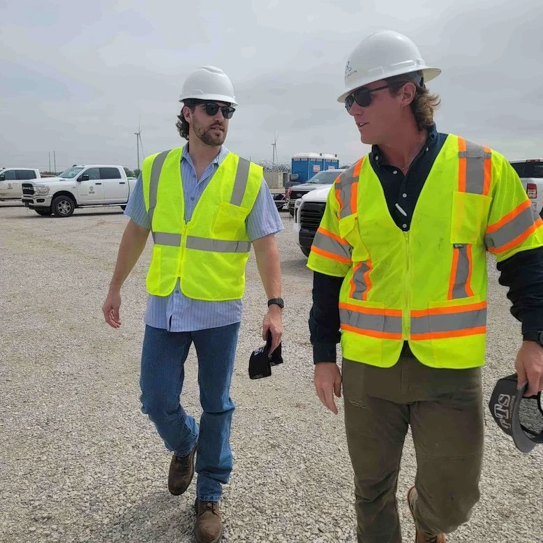 Two men wearing hard hats and high-visibility vests walking on a construction site with vehicles and wind turbines in the background.