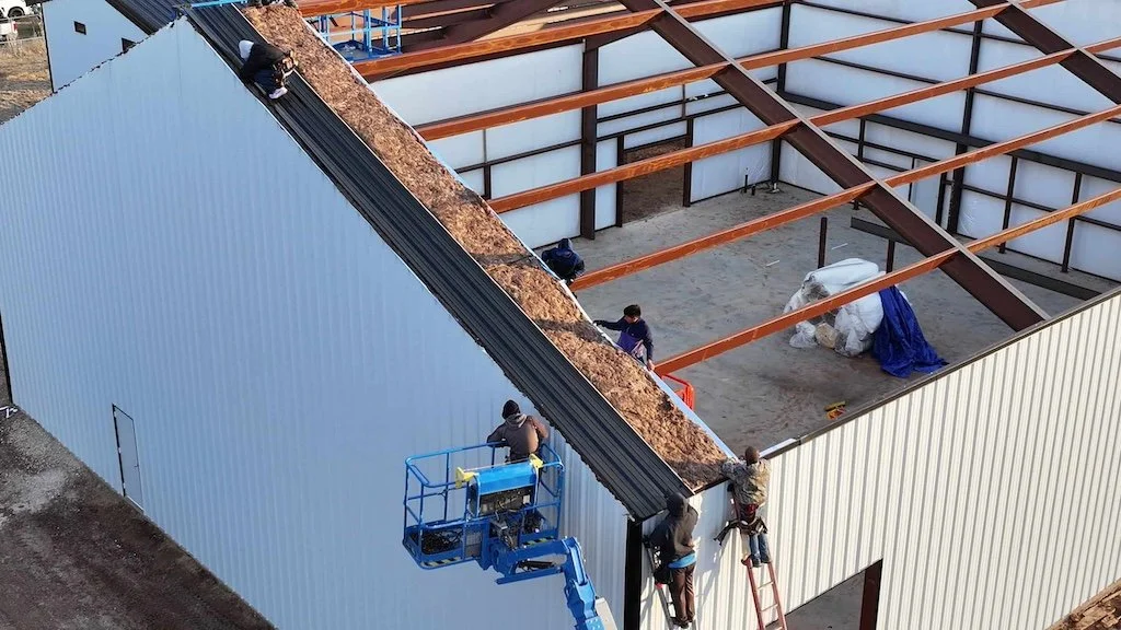 Construction workers on a building site, installing insulation and roofing on an industrial structure with exposed steel beams and a lift.