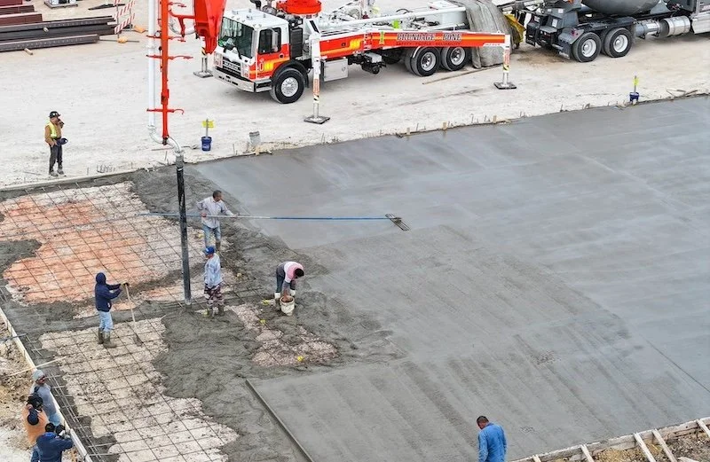 Construction site with workers laying and smoothing wet concrete on a large surface using tools and a concrete pump truck nearby.