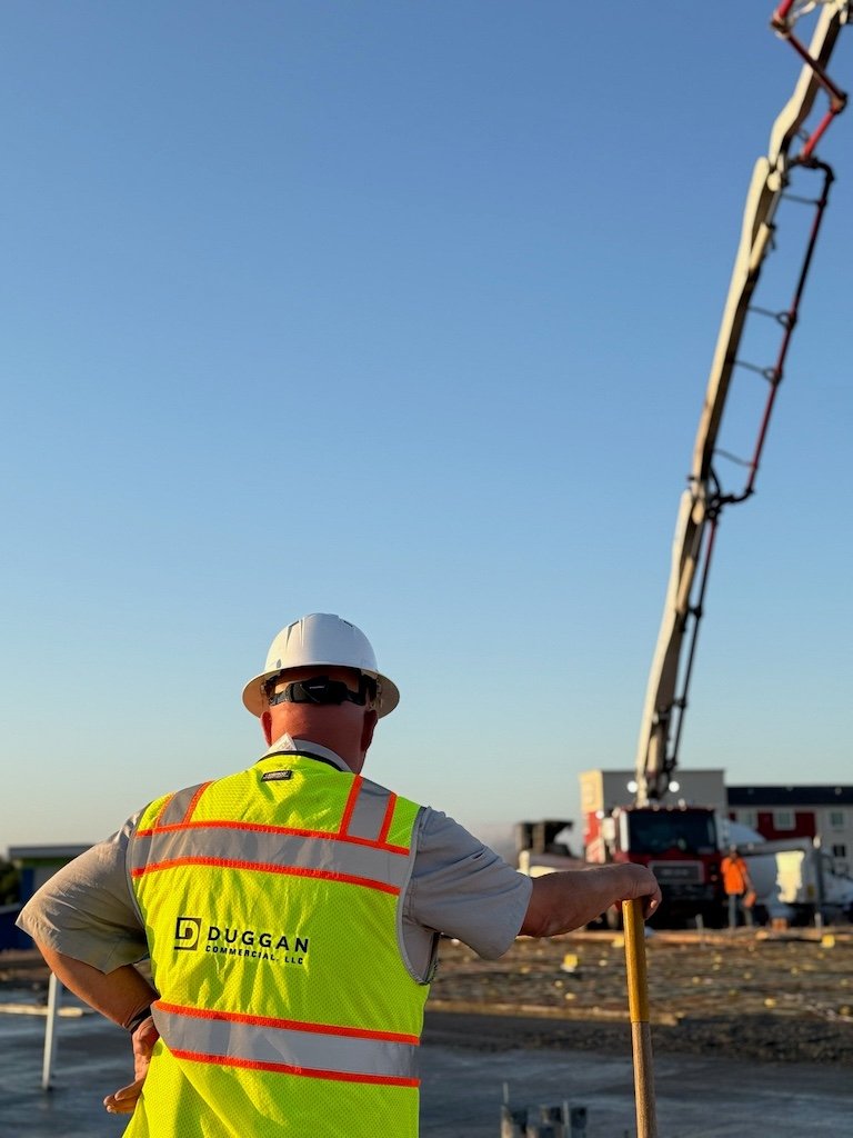 Construction worker in a safety vest and hard hat watches a concrete pump truck operate at a building site under a clear sky.