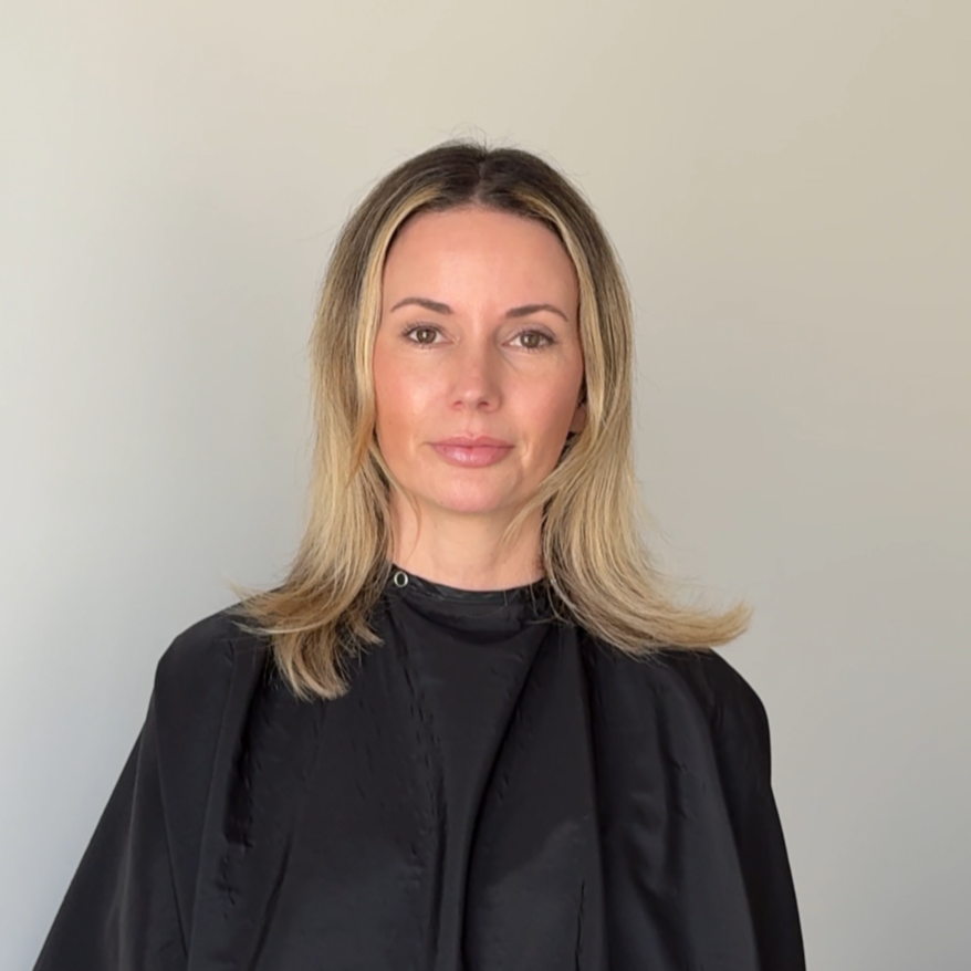 A woman in a black salon cape standing indoors against a plain wall.