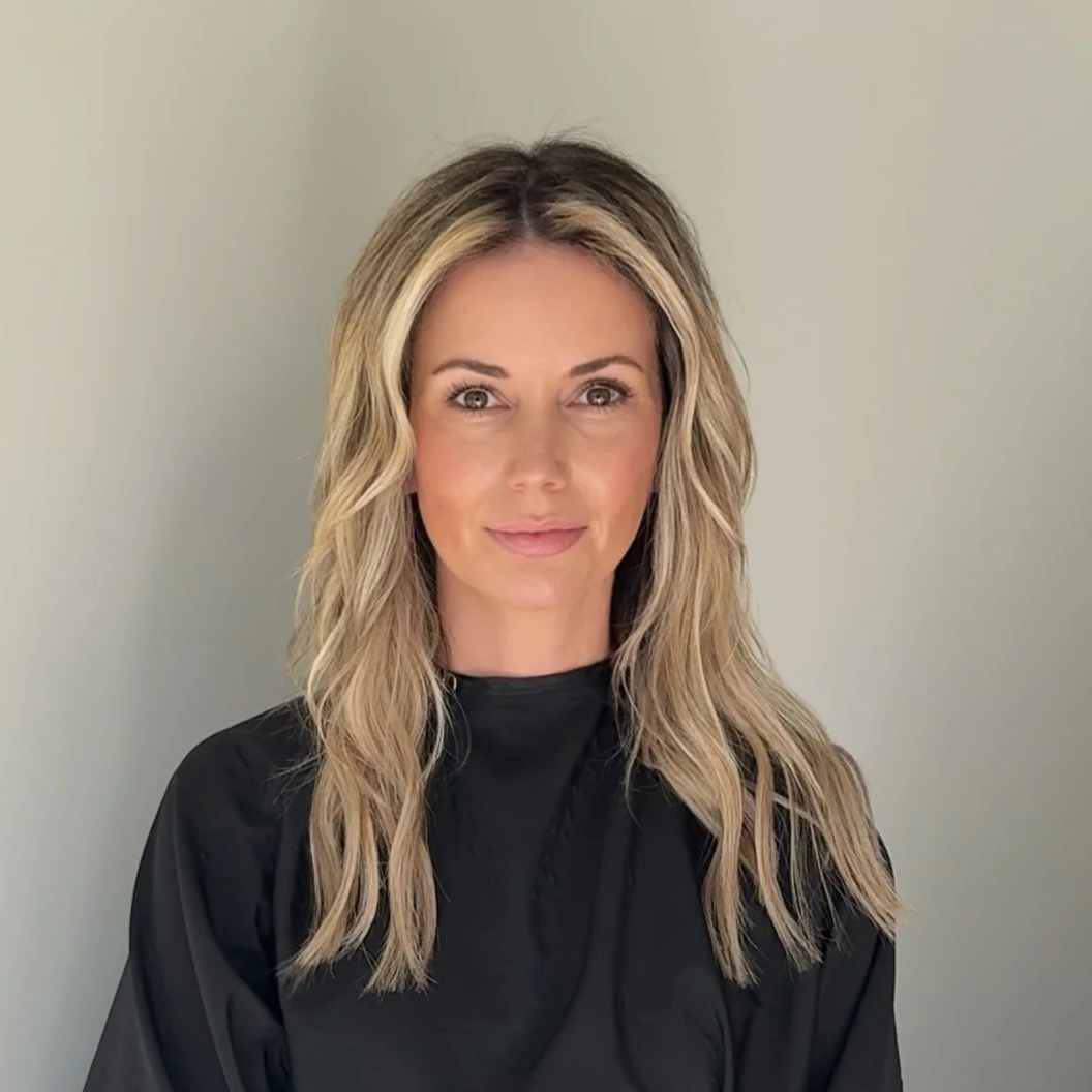 A woman with wavy blonde hair sitting against a plain wall, wearing a black cape typical for a salon or stylist appointment, and adorned with bracelets and a ring.