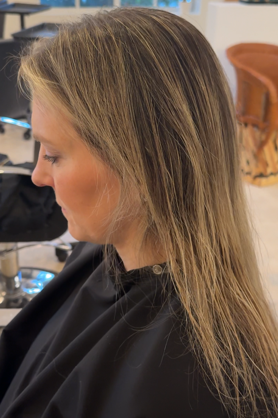Woman with blonde hair sitting in a hair salon chair, wearing a black cape, with hair wet and styled for a haircut or styling session. The salon has a large window showing trees outside, black chairs, a wooden armchair, and hair styling tools in the 