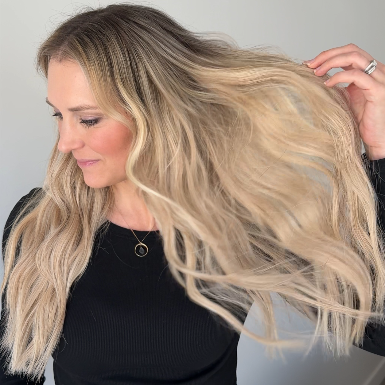 A woman with long wavy blonde hair wearing a black top and jeans, holding her hair to show its length and texture in a bright indoor setting.