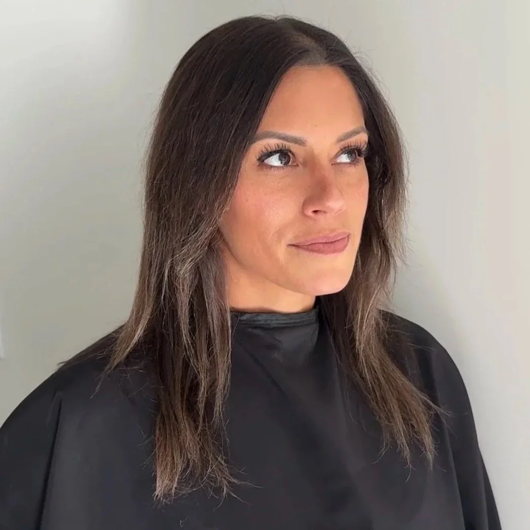 Woman with shoulder-length brown hair wearing a black salon cape, standing indoors against a light-colored wall.